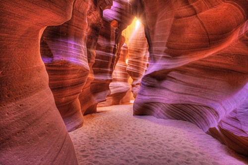 A narrow canyon with smooth, colorful rock walls and soft sand underfoot, illuminated by warm sunlight.