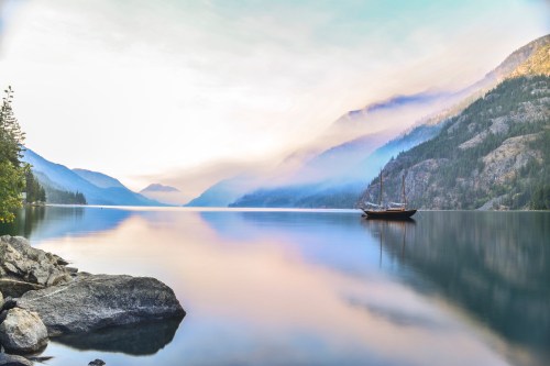 A serene lake surrounded by mountains, with a sailboat anchored and soft mist hovering over the water.