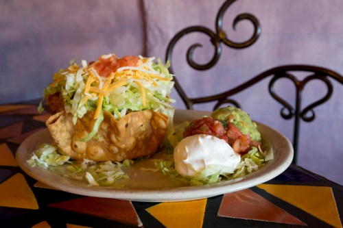 A colorful plate featuring a taco salad in a crispy shell, topped with lettuce, cheese, salsa, guacamole, and sour cream.