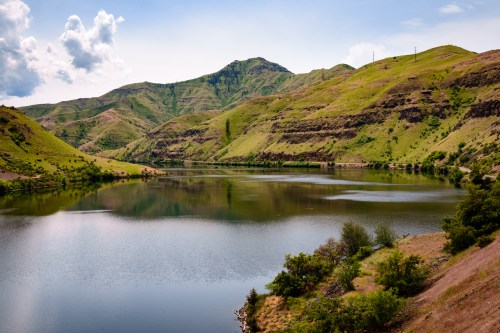 A serene lake surrounded by lush green hills under a partly cloudy sky. Reflections shimmer on the water's surface.