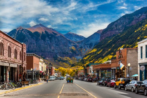 A scenic mountain town street lined with shops, colorful autumn trees, and towering mountains in the background.