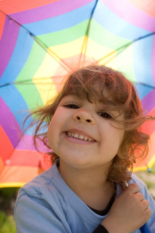 A smiling child with curly hair holds a colorful rainbow umbrella, enjoying a sunny day outdoors.