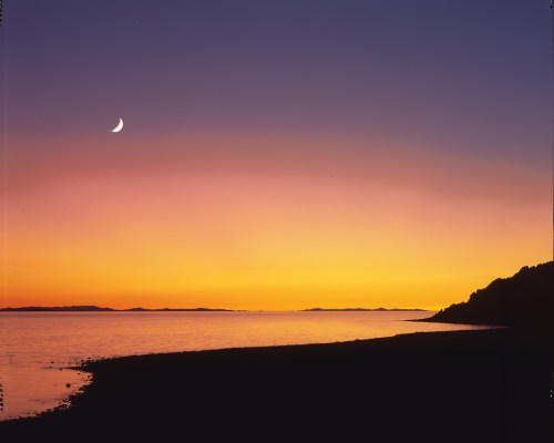 A serene sunset over a calm sea, with a crescent moon in the sky and silhouettes of distant islands on the horizon.
