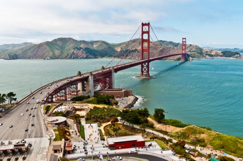 Aerial view of the Golden Gate Bridge spanning a bay, with hills in the background and people along the waterfront.