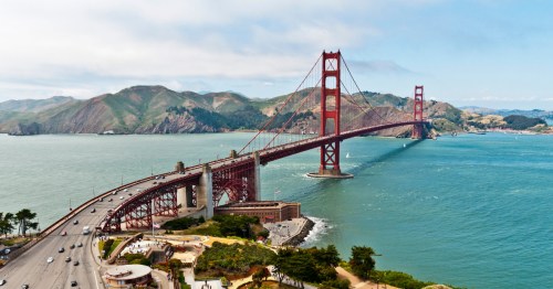 Aerial view of the Golden Gate Bridge spanning a bay, with hills in the background and vehicles on the roadway.