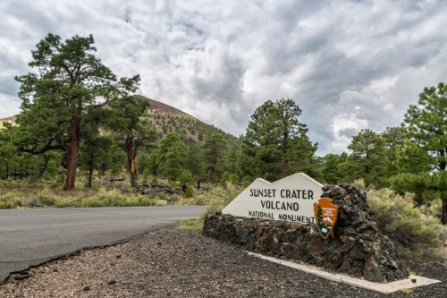 Sign for Sunset Crater Volcano National Monument, surrounded by trees and a cloudy sky. Road visible in the foreground.