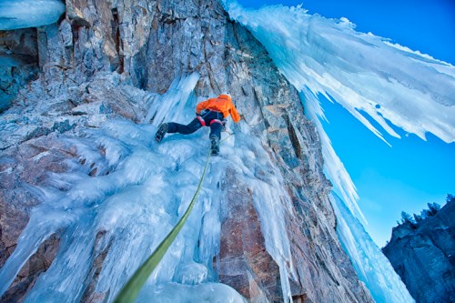 A climber ascends a frozen rock face, with icicles hanging above and a clear blue sky in the background.