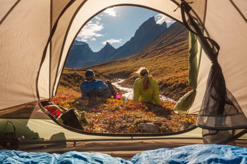 A view from inside a tent showing two people relaxing by a river in a mountainous landscape.