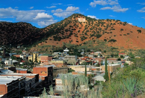 A scenic view of a small town nestled in a valley, surrounded by hills and blue skies with scattered clouds.