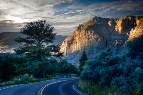 A winding road curves through a mountainous landscape, with trees and dramatic rock formations under a cloudy sky.