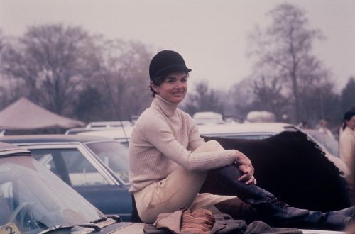 A woman in equestrian attire sits on a car hood, smiling, with a misty background of parked vehicles and trees.