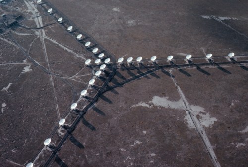 Aerial view of satellite dishes arranged in a grid on a barren landscape, casting long shadows.