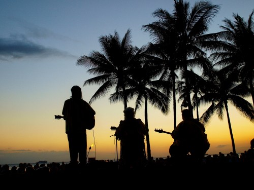 Silhouettes of musicians playing guitars against a sunset backdrop with palm trees.