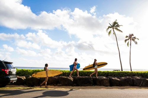 Three surfers walk along a rocky path by the beach, carrying surfboards under a sunny sky with palm trees.