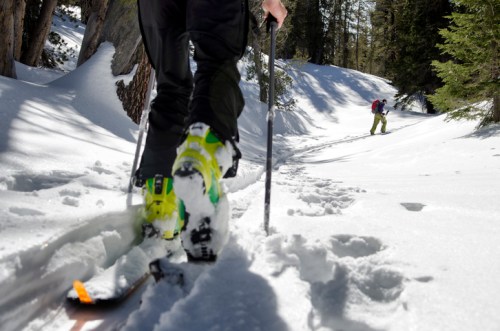 A skier ascends a snowy trail, using poles, with another skier visible in the background among trees.