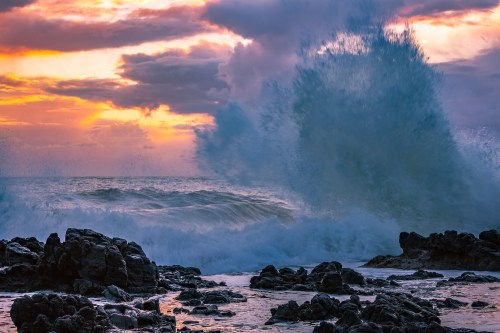 Waves crash against rocky shore at sunset, with vibrant colors in the sky and dramatic cloud formations.