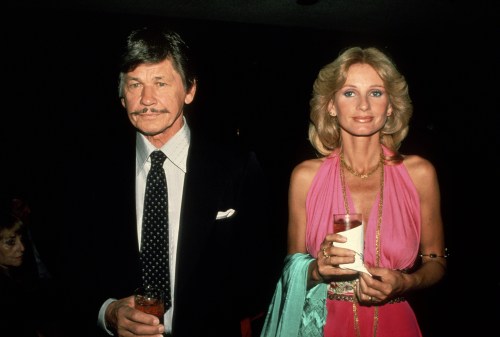 A man in a black suit and a woman in a pink dress hold drinks at a formal event, smiling for the camera.