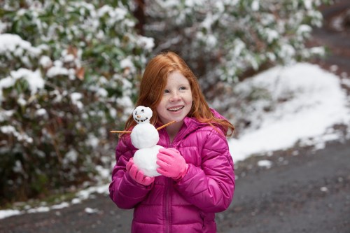 A smiling girl with red hair in a pink jacket holds a small snowman in a snowy outdoor setting.