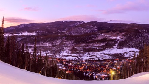 A scenic winter landscape at dusk, featuring snow-covered mountains and a small town illuminated in the valley below.