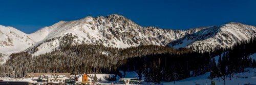 Snow-covered mountains rise against a clear blue sky, with a small village nestled at the base among evergreen trees.