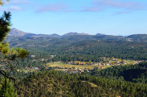 A panoramic view of a small town surrounded by lush green forests and mountains under a clear blue sky.