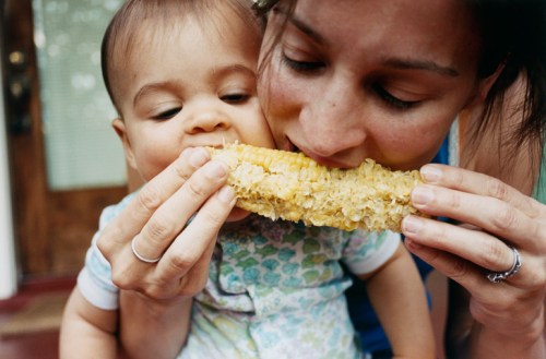 A woman and a baby share a corn on the cob, both taking bites from opposite ends, smiling and enjoying the moment.