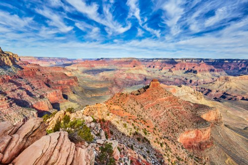 Vast view of the Grand Canyon with colorful rock formations and a blue sky filled with wispy clouds.
