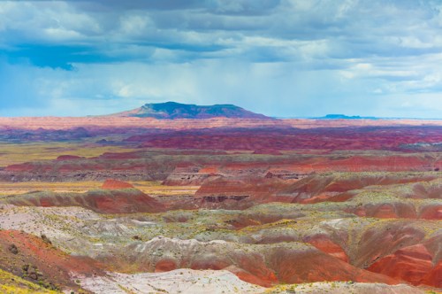 Vast landscape of colorful, layered hills under a cloudy sky, showcasing shades of red, orange, and green.