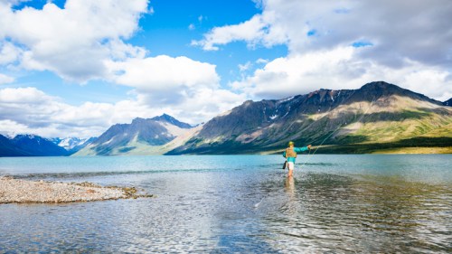 A person fishing in a serene lake surrounded by mountains and blue skies, with clouds scattered above.