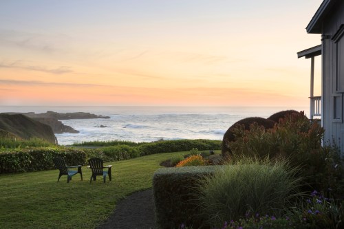 A serene coastal view at sunset, featuring two chairs on a grassy lawn overlooking the ocean.