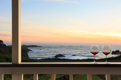 Two glasses of red wine sit on a balcony railing, overlooking a serene ocean sunset.