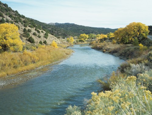 A serene river flows through a landscape of autumn trees and rolling hills, showcasing vibrant yellow foliage.