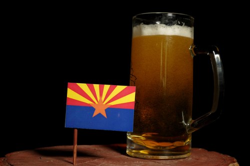 A large mug of beer next to a small Arizona state flag on a wooden surface against a black background.