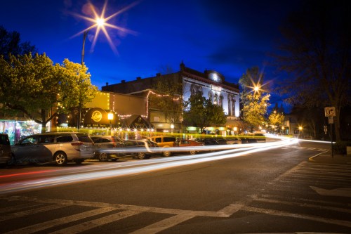 A vibrant street scene at night, featuring light trails from cars, trees, and a historic building illuminated by streetlights.
