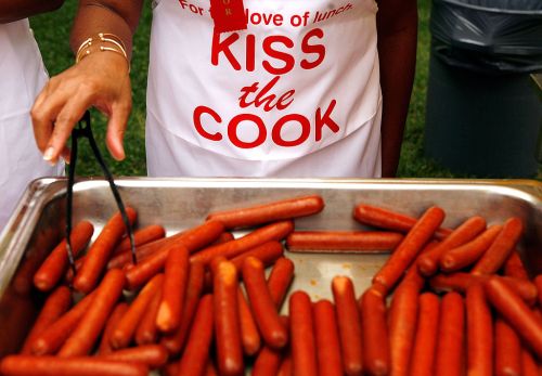 A person in an apron reading "Kiss the Cook" serves hot dogs from a tray at an outdoor event.