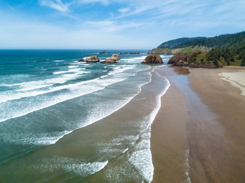 Aerial view of a sandy beach with gentle waves, rocky formations, and lush green hills under a blue sky.