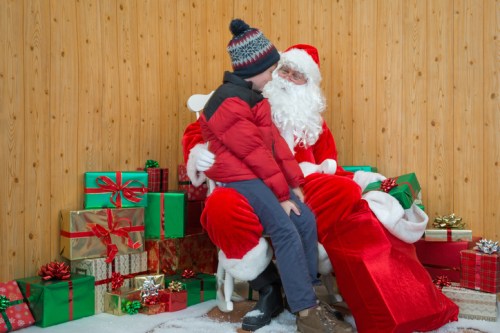 A child sits on Santa's lap in front of a backdrop of colorful Christmas presents.