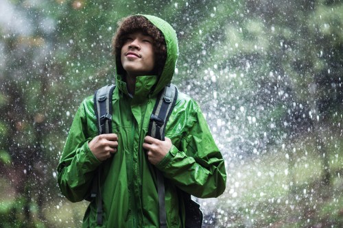 A young person in a green rain jacket stands outdoors, looking up with a smile as rain falls around them.