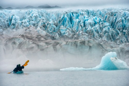 A kayaker paddles near a massive glacier, surrounded by icy blue hues and misty mountains in the background.