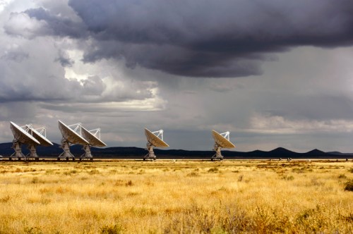 A row of large satellite dishes in a grassy field under a cloudy sky, with distant mountains in the background.