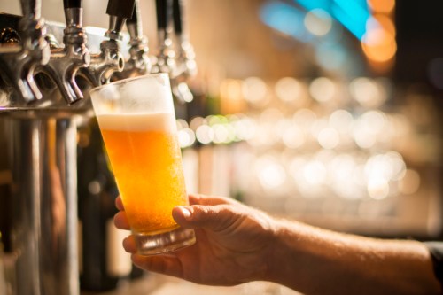 A hand holds a glass of beer, with a tap system and blurred lights in the background.