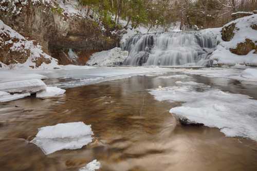 A serene winter scene featuring a frozen waterfall surrounded by snow and ice, with a calm river in the foreground.