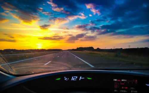 View from a car dashboard at sunset, showing a highway and colorful sky with clouds. Speedometer reads 112 km/h.