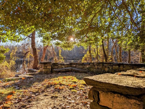 A serene outdoor scene with a stone wall, trees, and sunlight filtering through leaves by a calm water body.
