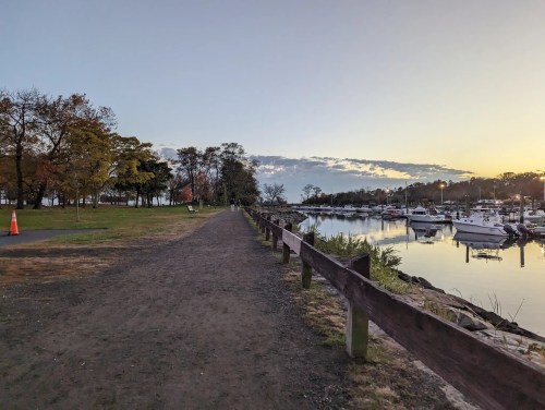 A peaceful waterfront path lined with trees, leading to a marina with boats at sunset.