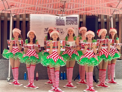A group of young performers in festive costumes singing under a tent, wearing Santa hats and striped stockings.