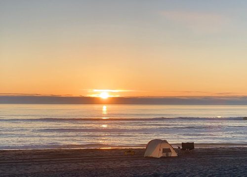 A serene beach at sunset, featuring a tent and a chair on the sand with calm waves in the background.