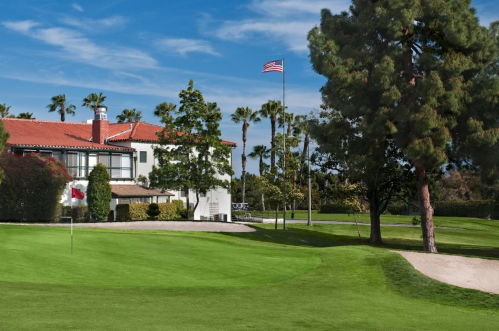 A golf course with a flag, palm trees, and a white building under a blue sky with an American flag flying.