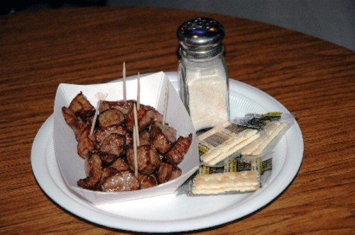 A plate with skewered meat pieces, salt shaker, and packets of crackers on a wooden table.