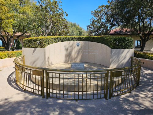 A circular stone memorial surrounded by a gold railing, set in a landscaped area with trees and blue sky.